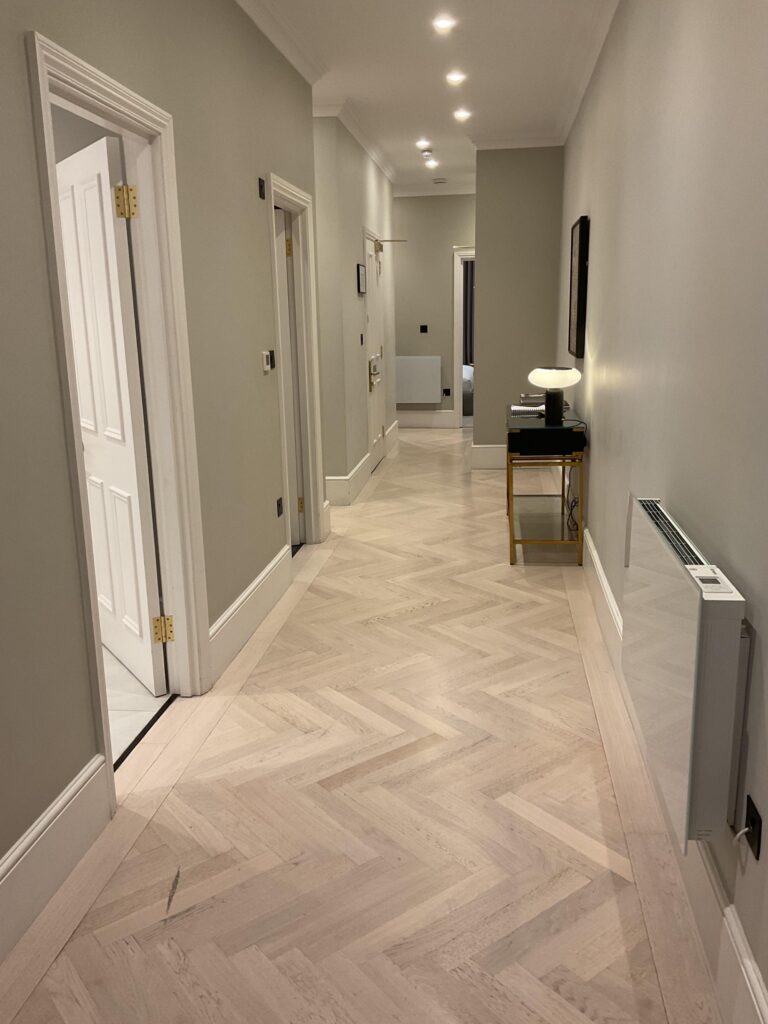 Long corridor with pale herringbone flooring, recessed downlights and brass door furniture