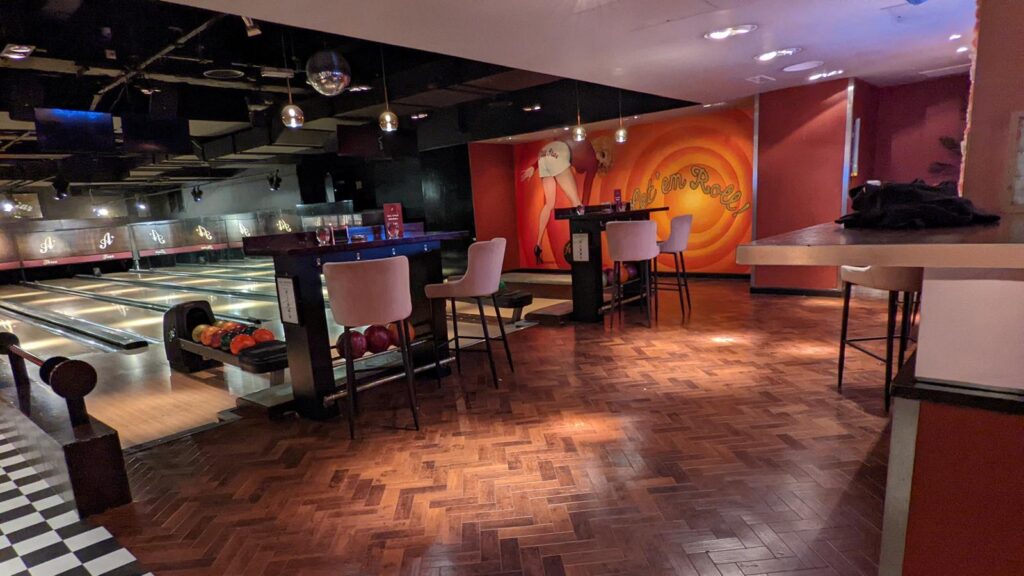 Bowling lanes beside a lounge with herringbone timber floor, bar tables and a large mural